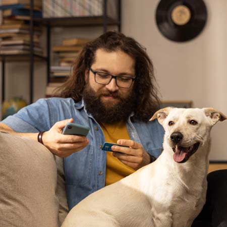 Long haired man with beard and glasses, sitting on couch with white dog with phone and credit card.