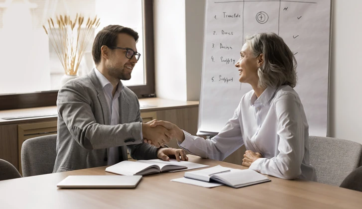Happy mature businesswoman giving handshake to younger colleagues man, discussing agreement at meeting table. Employer and candidate shaking hands after successful job interview