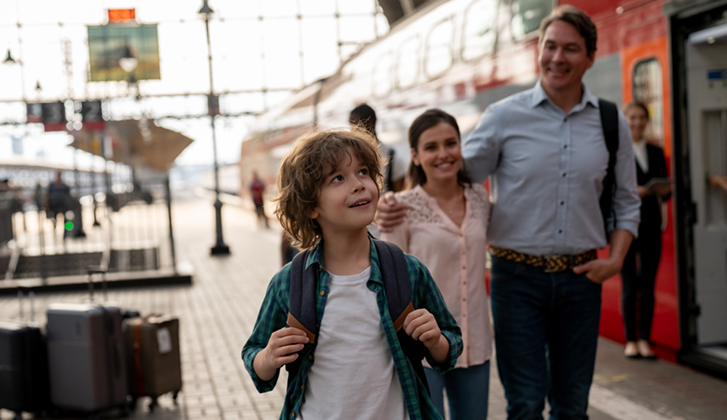 Child with long curly hair holding backpack staring off screen in wonder with parents looking proudly at them in the background at a train station