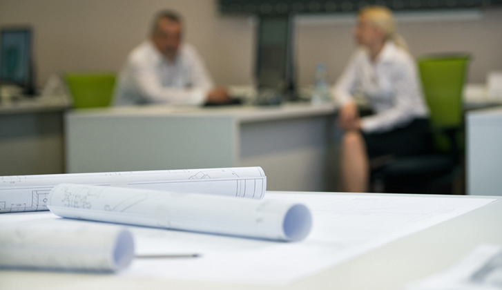 Close-up image of rolled blueprints with an out-of-focus of man and woman in business attire sitting at a desk in the background