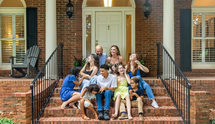 Blended family sitting together happily on the front steps to their large brick home.