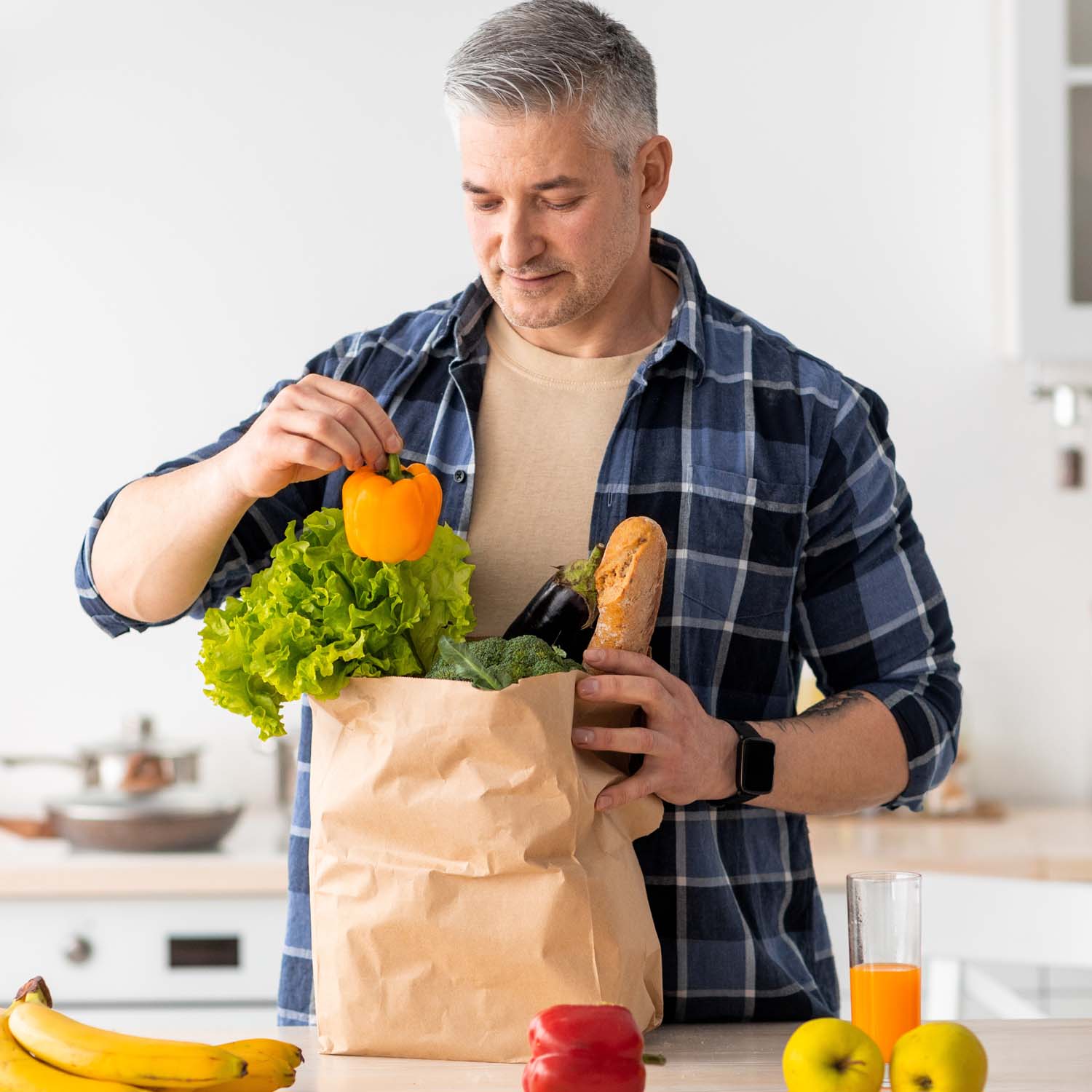 Older man in plaid shirt emptying his grocery bag full of fruits and vegetables in his kitchen.