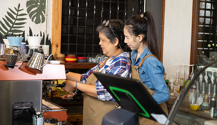 Young barista female hiring senior, elderly, 60s pensioner worker working in cafe bakery small business shop, two waitresses women standing behind coffee counter using technology cash register machine