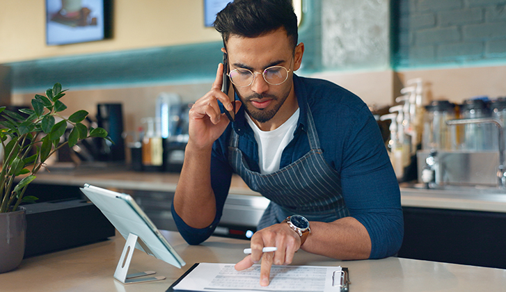 A business owner is on a phone call while reviewing paperwork in a restaurant or café setting. The scene includes a supply chain checklist or inventory documents..